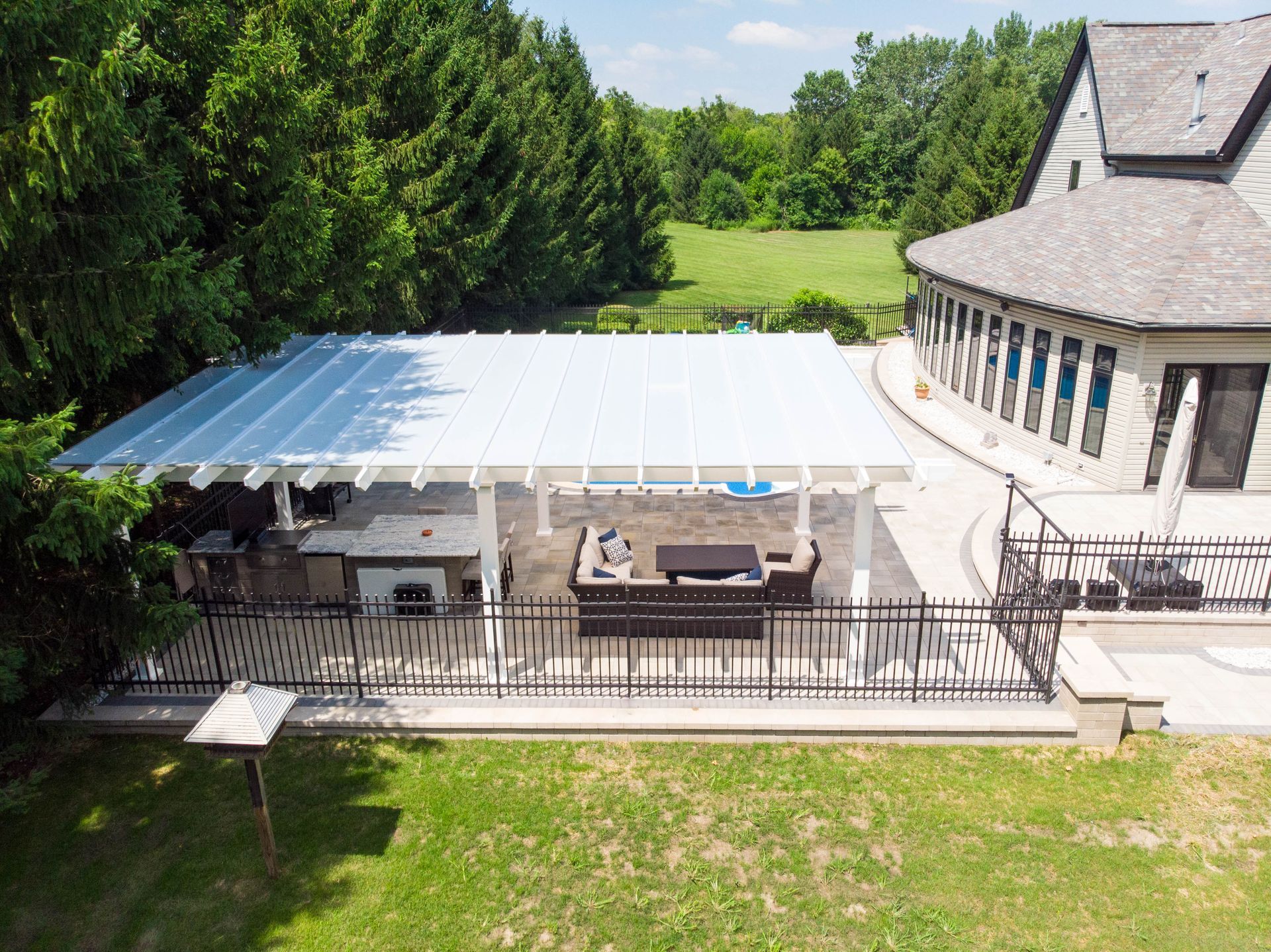 An aerial view of a white patio pergola with lounge furniture next to a suburban house and a grassy yard.