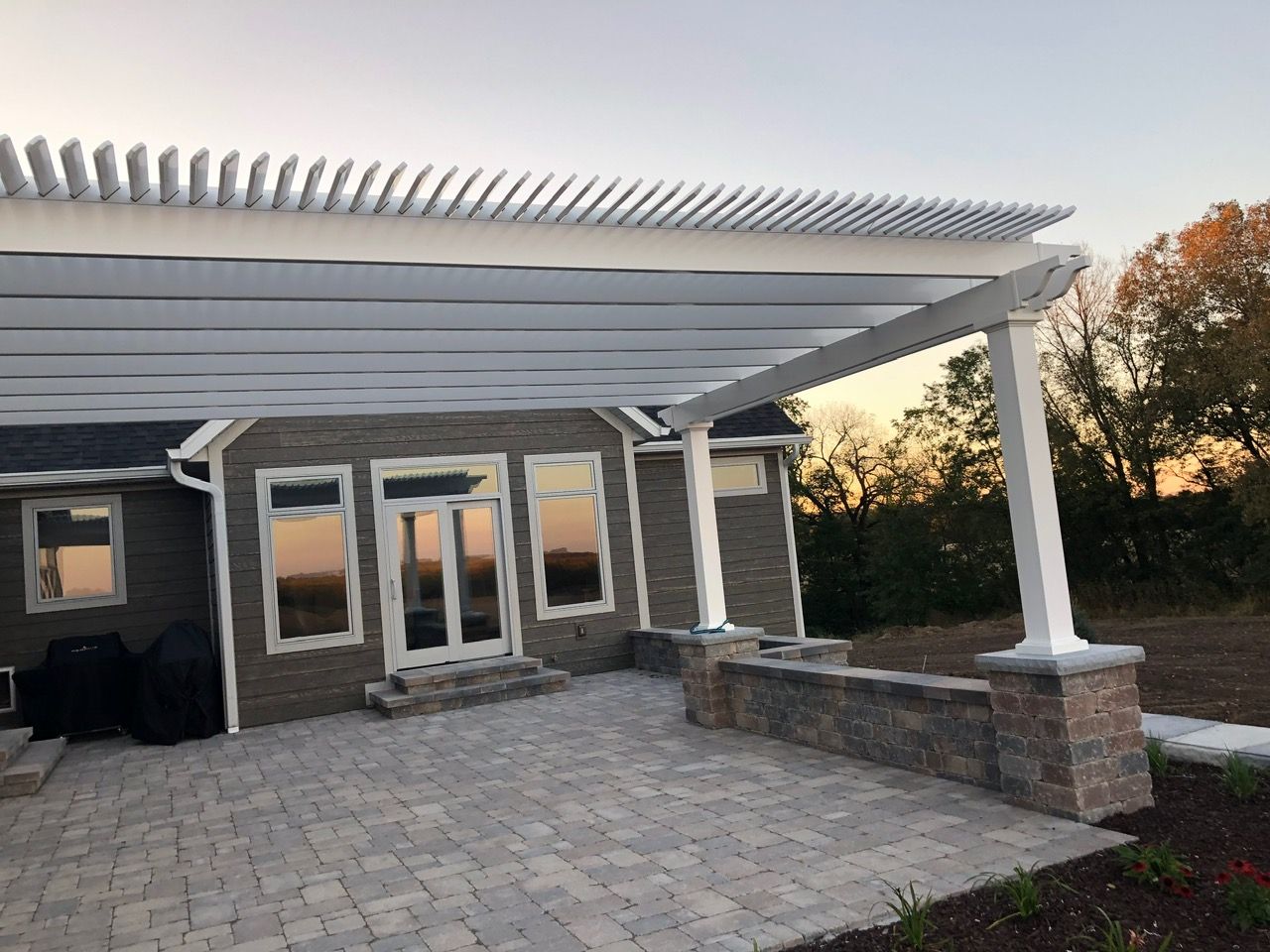 A patio with a white pergola featuring a retractable fabric shade, attached to a brick house with stone walls at sunset.