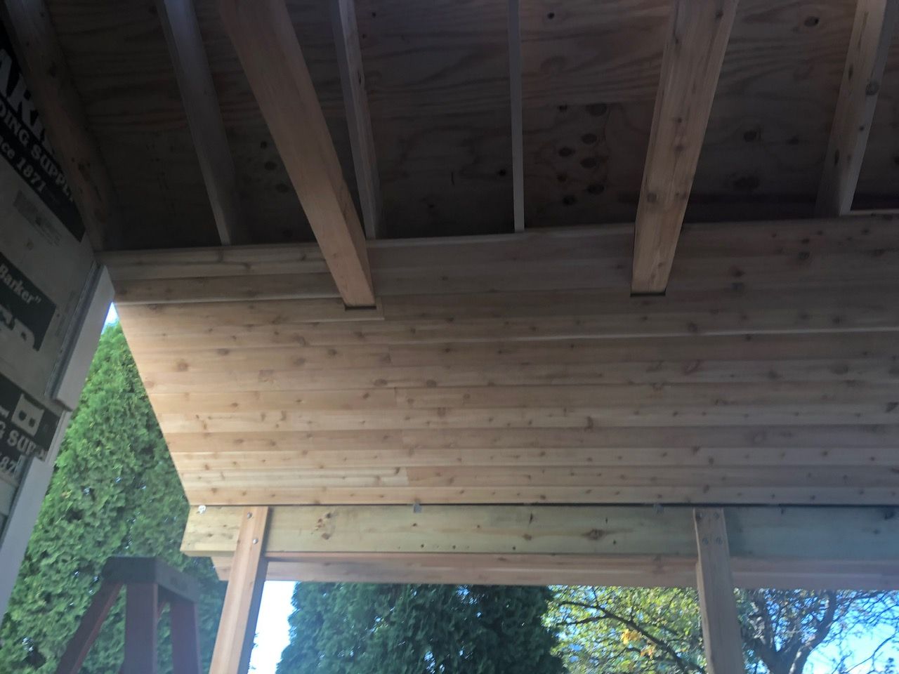 Construction of a wooden porch ceiling with exposed joists, light-colored wooden planks, and green trees in the background.