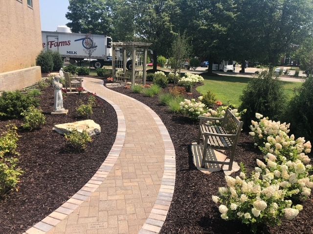 A winding paver walkway leads through a mulched garden with white flowering shrubs, a stone bench, and a trellis.