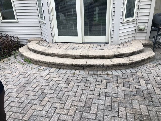 Two curved, light-colored paver steps leading to glass double doors set against a light-gray vinyl siding exterior.