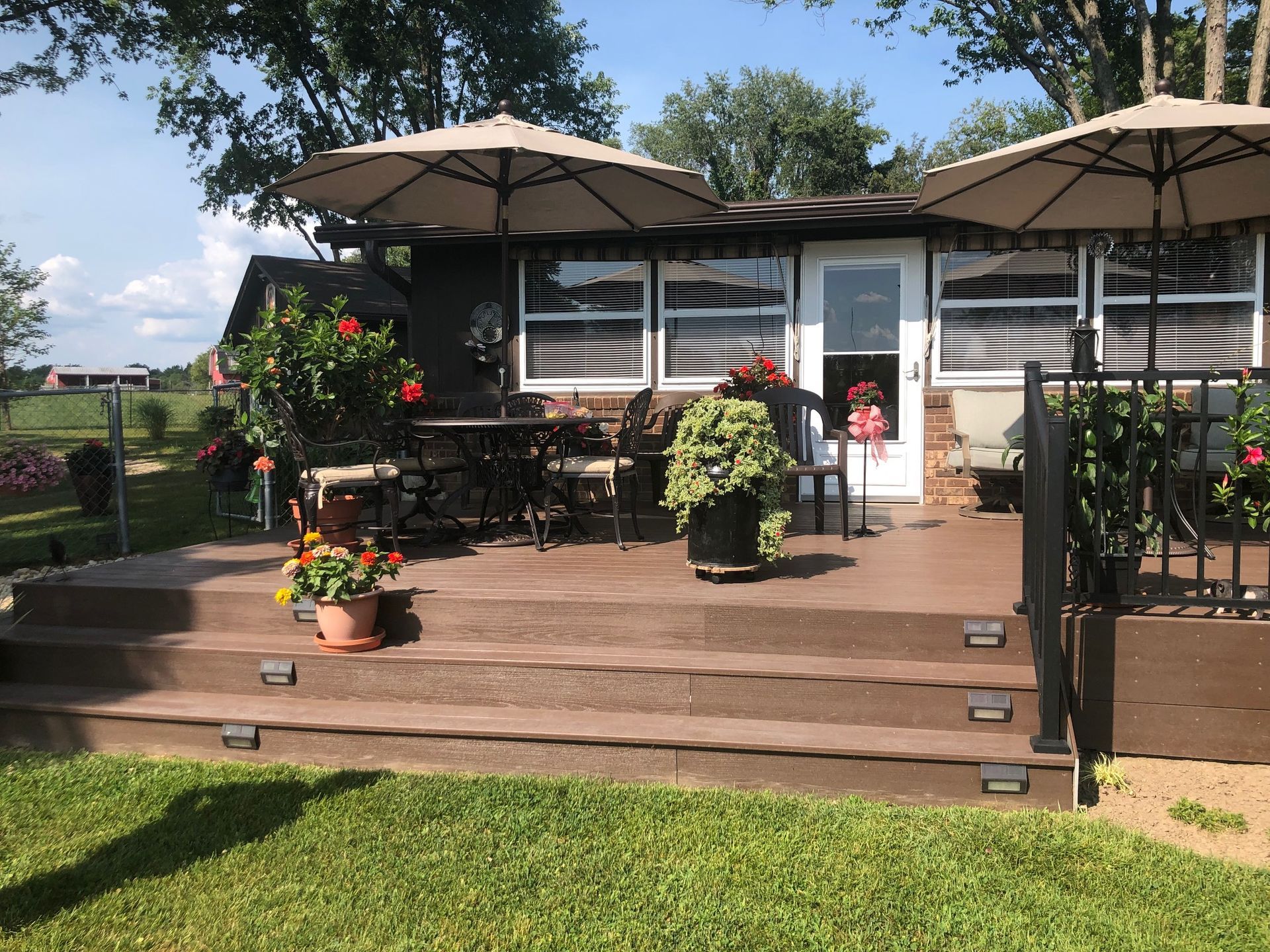 A dark brown wooden deck with two umbrellas, outdoor furniture, and potted plants in front of a house on a sunny day.