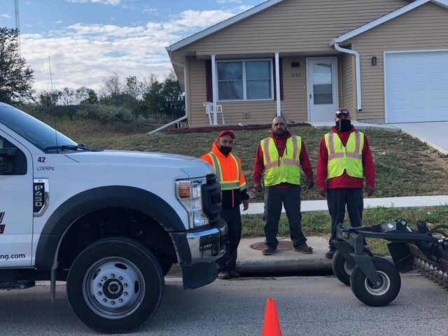 Three workers in high-visibility vests stand next to a utility truck and construction equipment in a residential street.