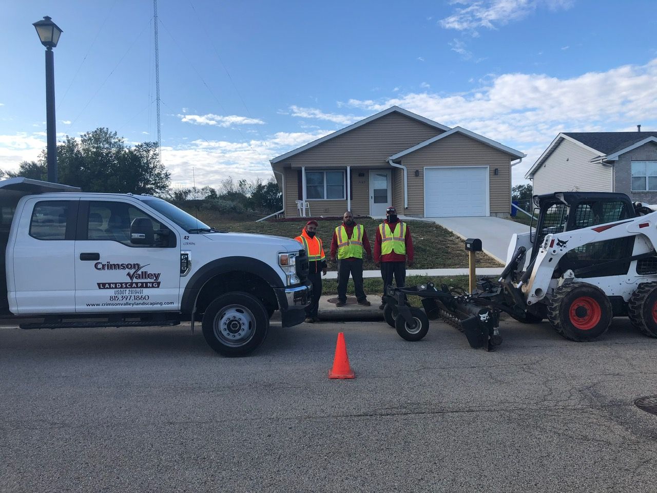 Three workers in high-visibility vests stand next to a white service truck and a white skid-steer loader on a street.