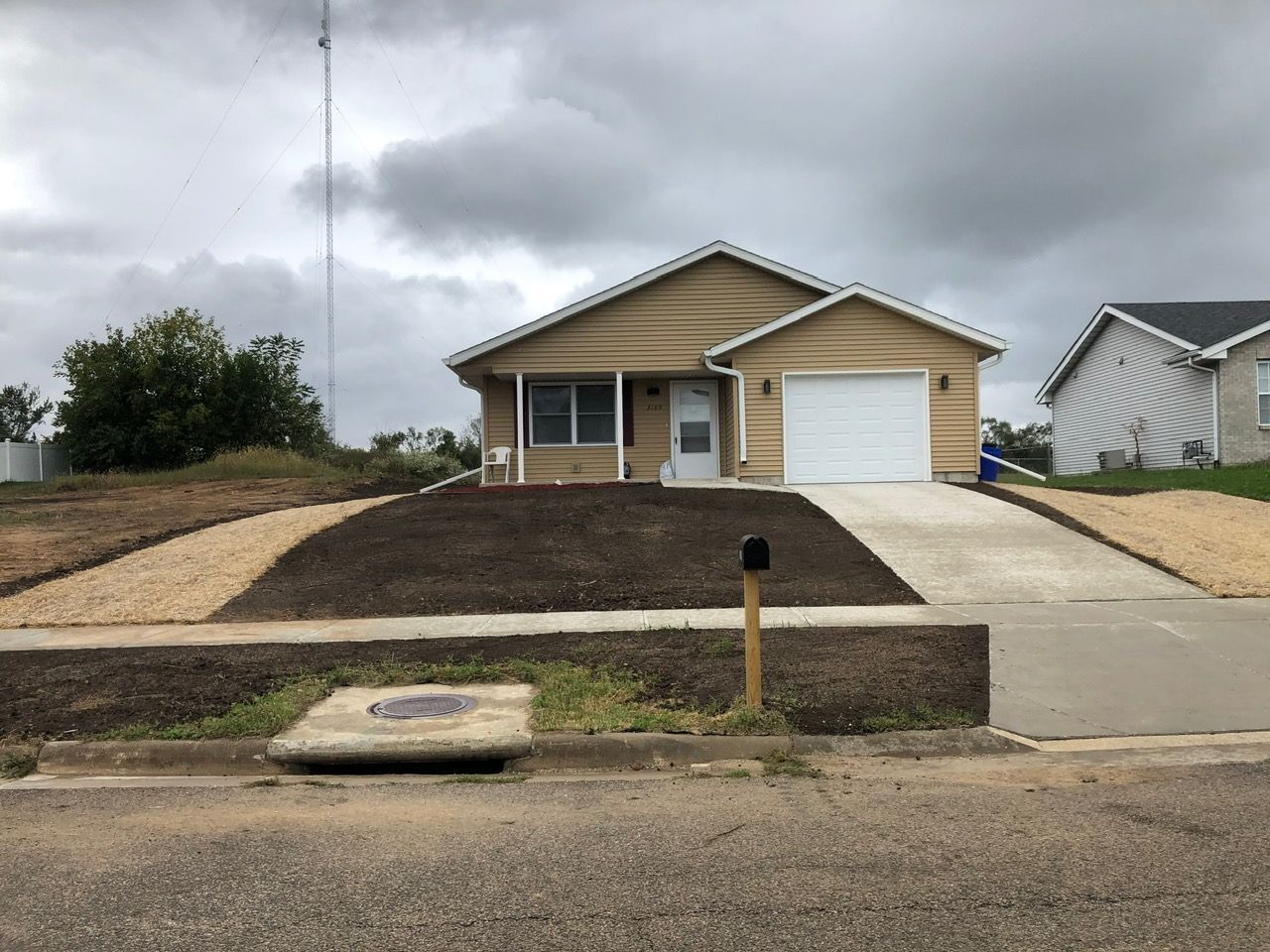 A tan, one-story house with a white garage door, a gravel walkway, and a front yard recently prepped with dark soil.