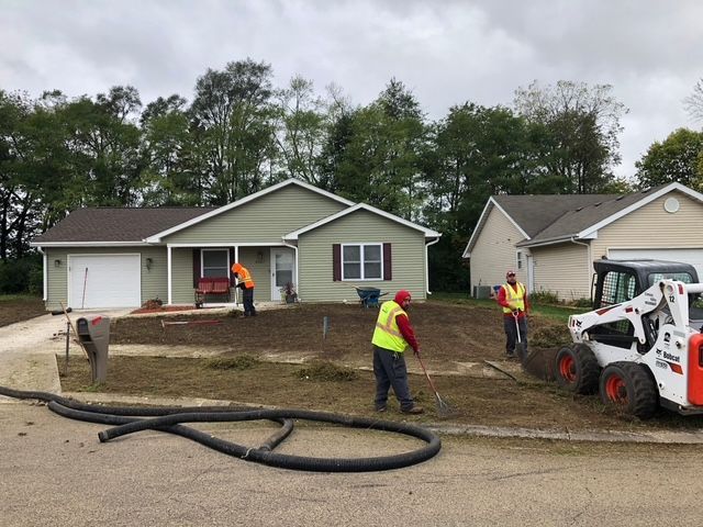 Three workers in high-visibility vests landscaping the front yard of a light green house with a white garage.