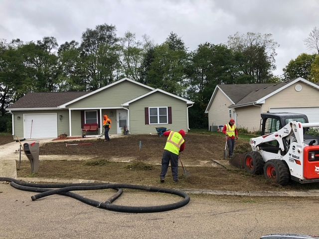 Construction workers in safety vests perform landscaping work with a skid steer in front of a green suburban house.