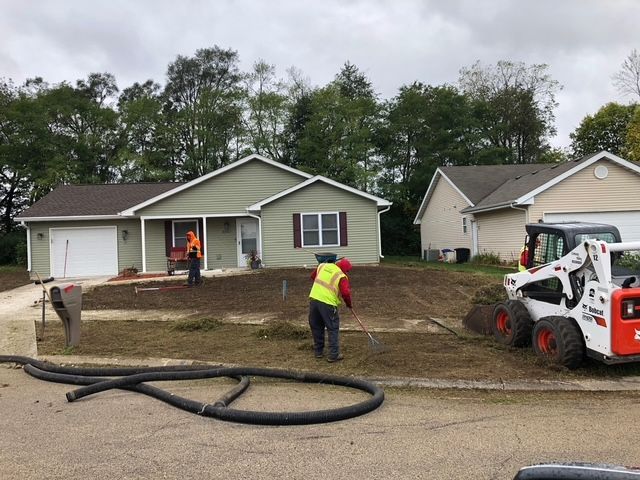 Construction workers in high-visibility vests work on a residential yard with a skid steer and heavy equipment.