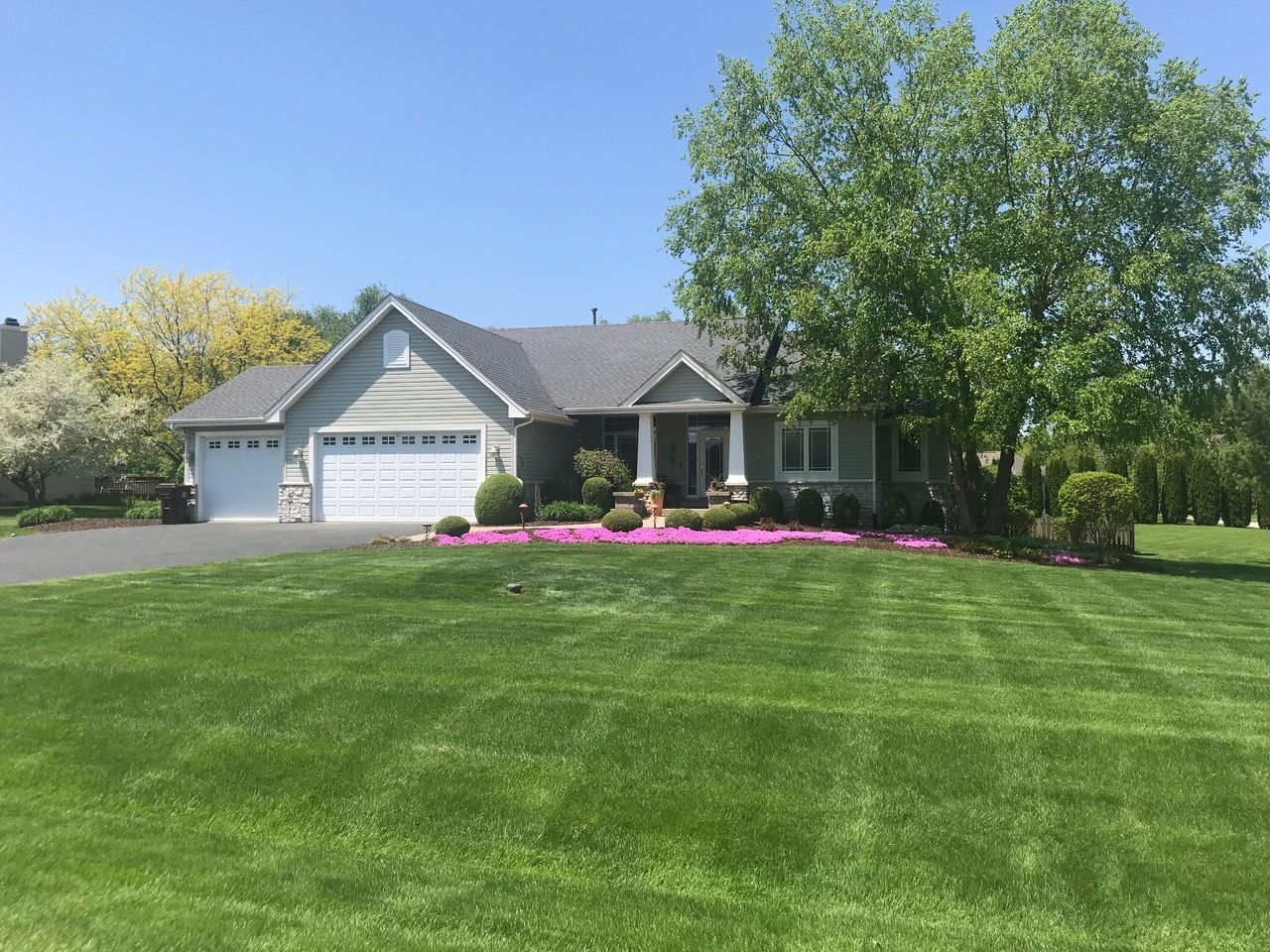 A grey-roofed single-story suburban house with a large green lawn, two-car garage, and a patch of vibrant pink flowers.