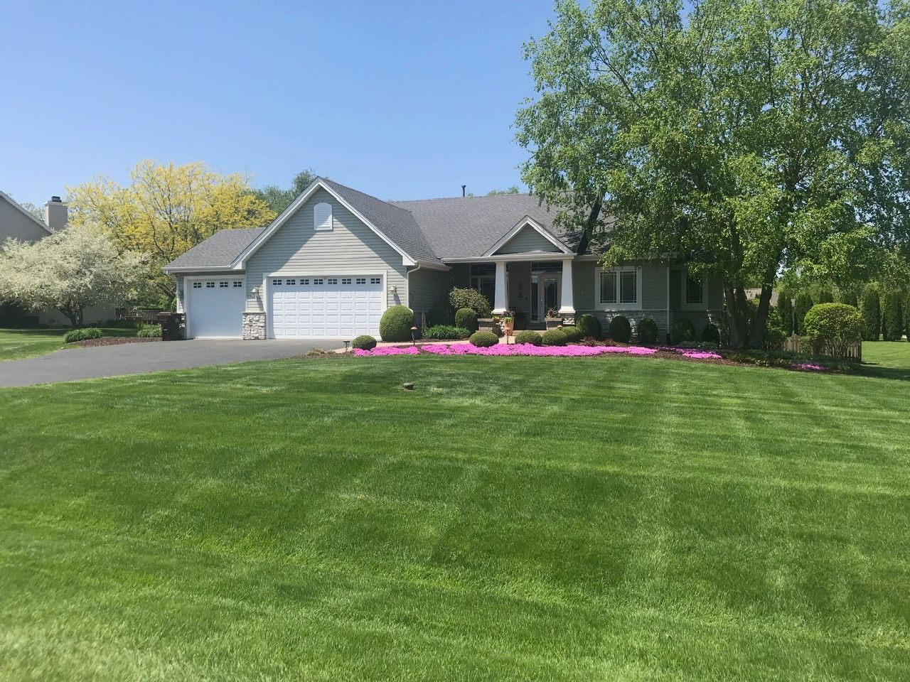 A one-story suburban house with a two-car garage, a manicured lawn, and a vibrant bed of pink flowers under large trees.