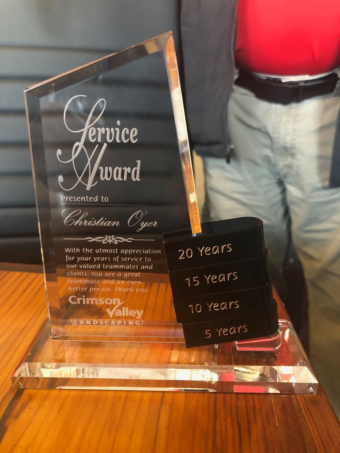 A glass Crimson Valley Service Award plaque for 20 years of service presented to Christian Oyan, sitting on a wooden table.