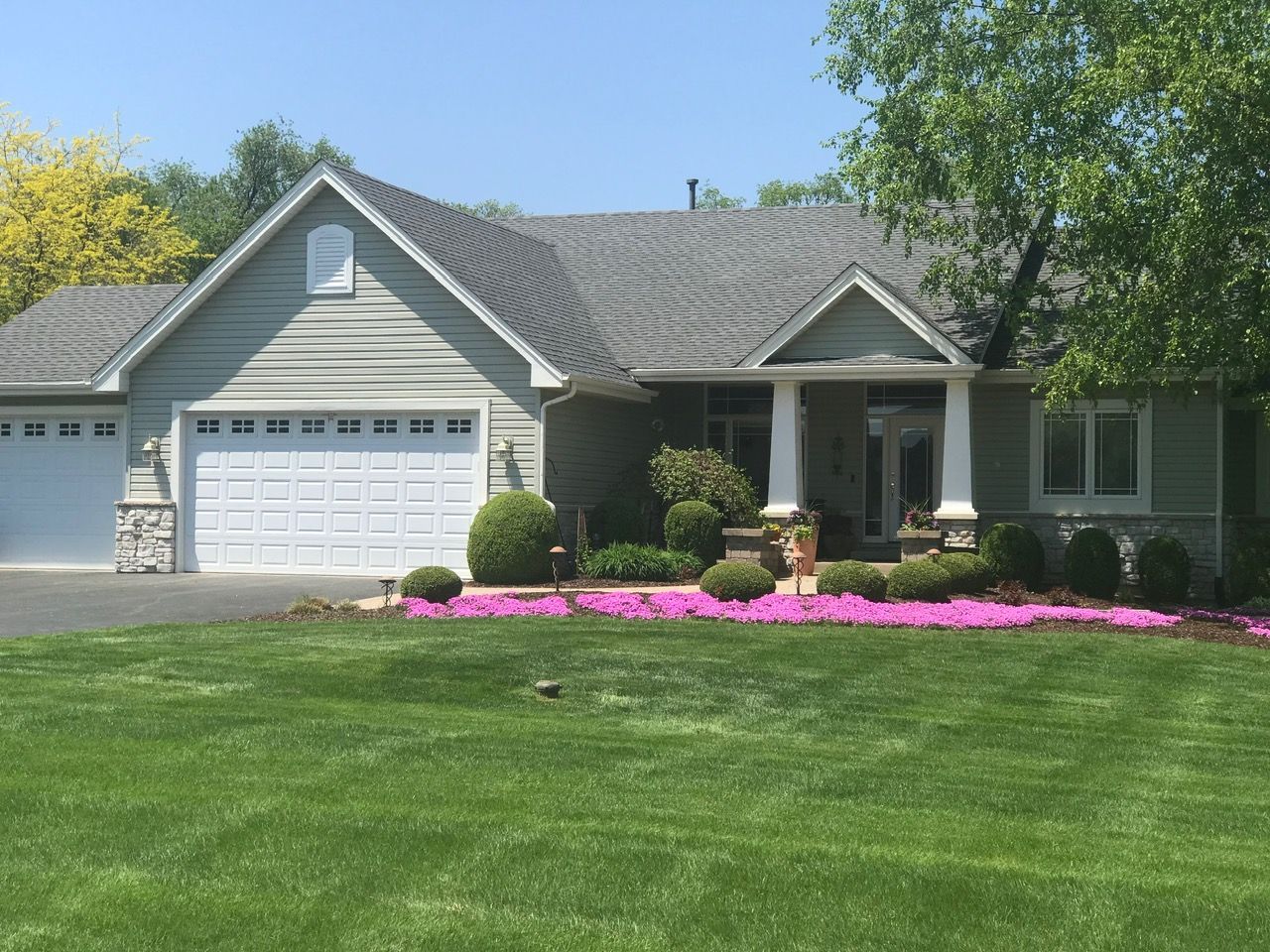A suburban house with a pale green exterior, gray shingled roof, a two-car garage, and a lawn with bright pink flowers.