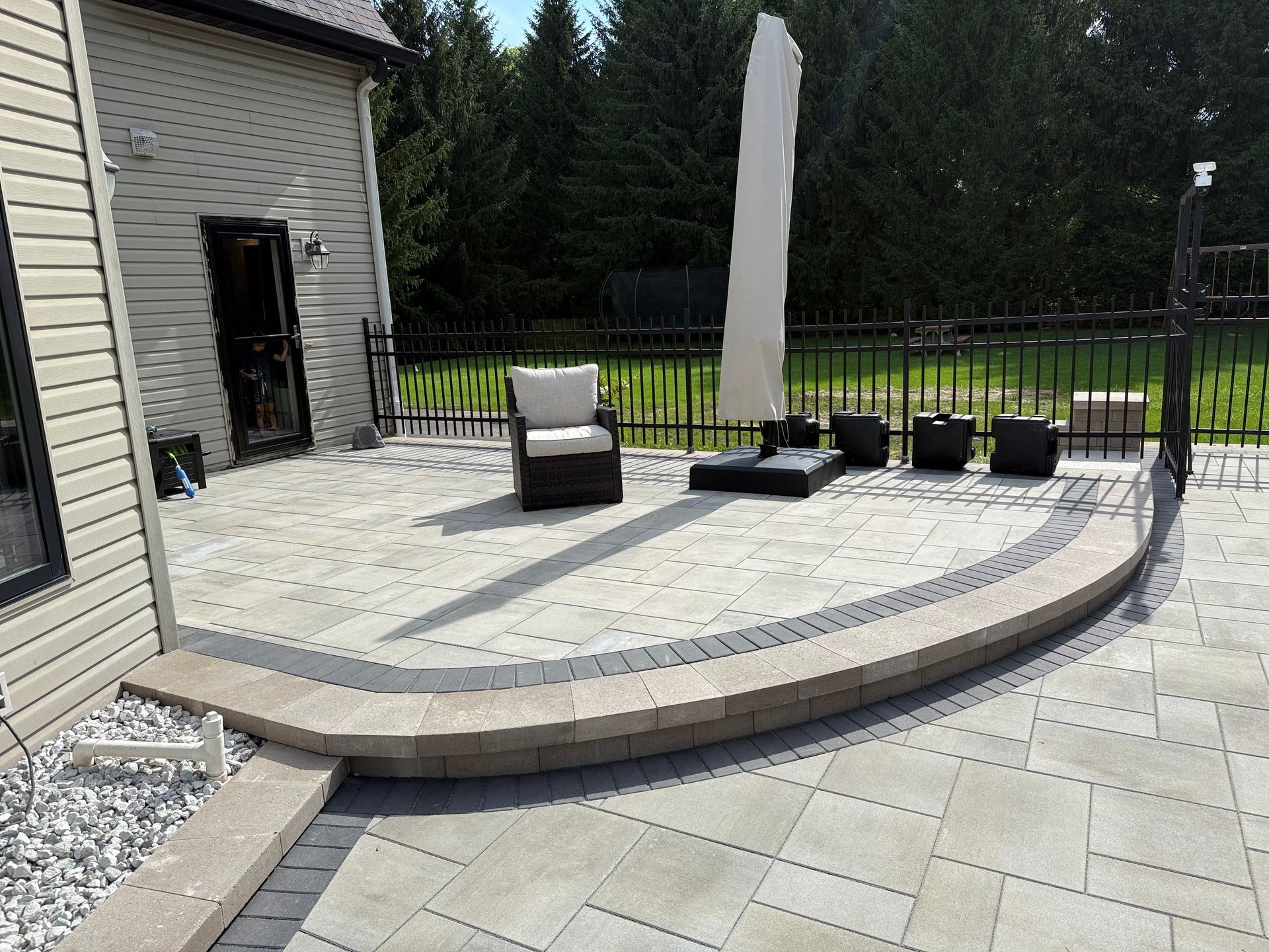 A backyard patio with light-colored pavers, a curved retaining wall, a patio chair, and a closed umbrella.