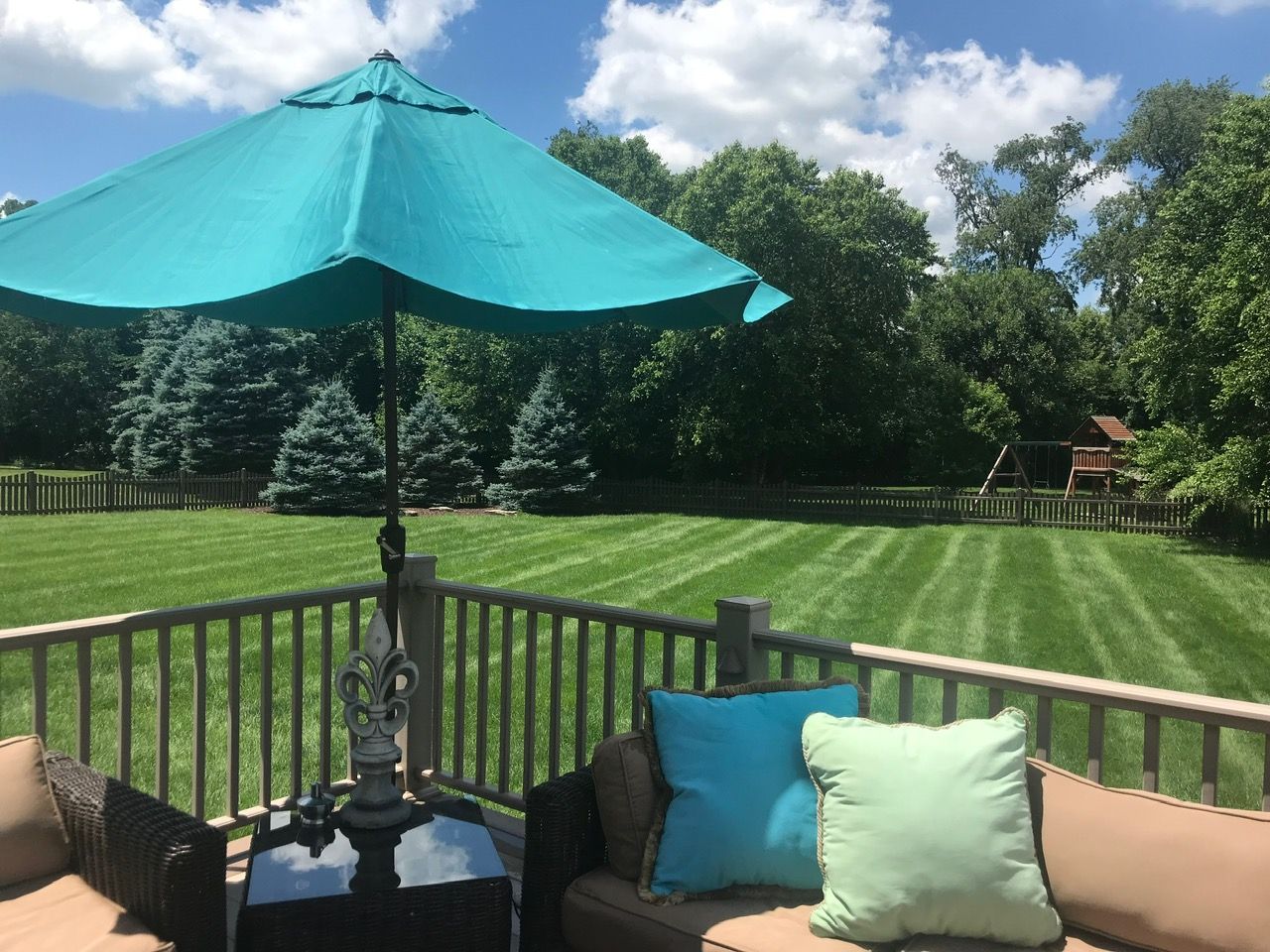 A turquoise umbrella over outdoor patio furniture on a deck, overlooking a mowed grassy lawn and trees under a blue sky.