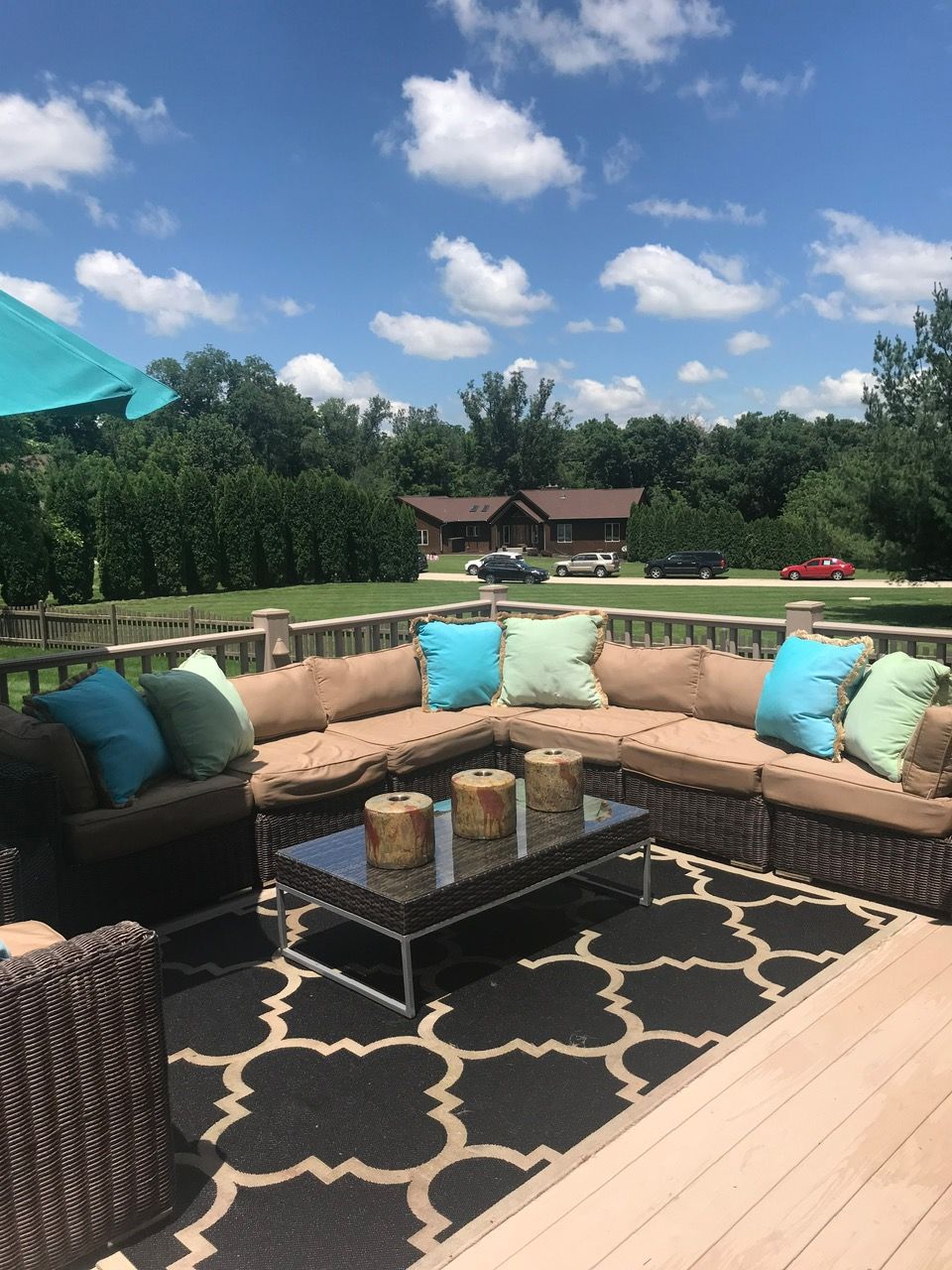 A patio with a brown wicker sectional, beige cushions, teal pillows, and a glass coffee table on a patterned rug.