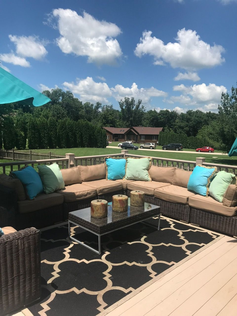 Outdoor patio seating with a wicker sectional, tan cushions, colorful throw pillows, and a glass-top table on a deck.
