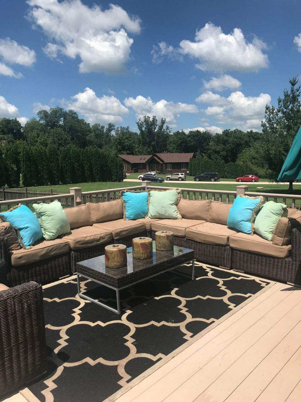 Outdoor wicker sectional sofa with blue and green pillows and a glass-top coffee table on a deck with a patterned rug.