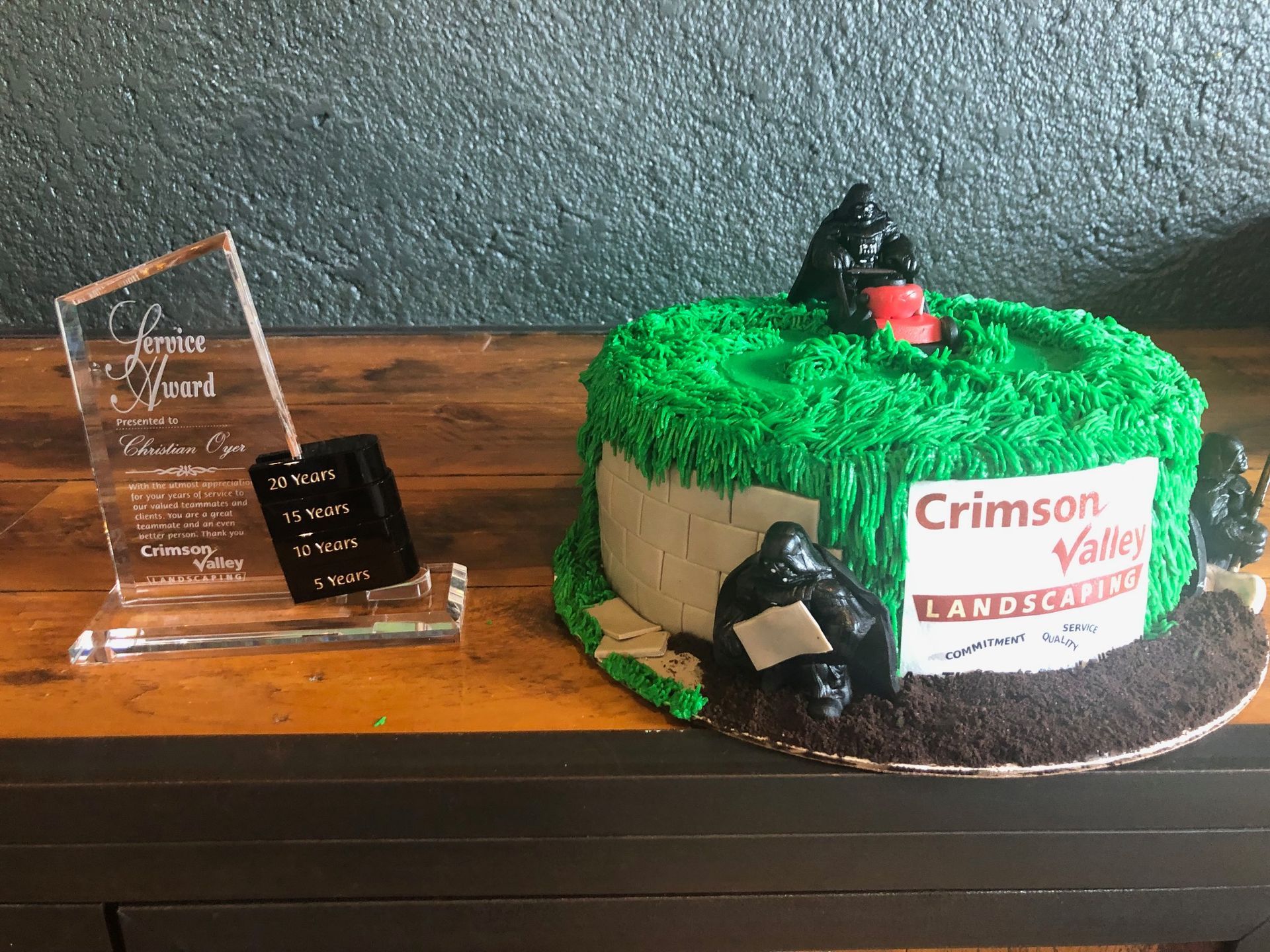 An award plaque next to a green-frosted cake with a Crimson Valley Landscaping sign, topped with small toy figurines.