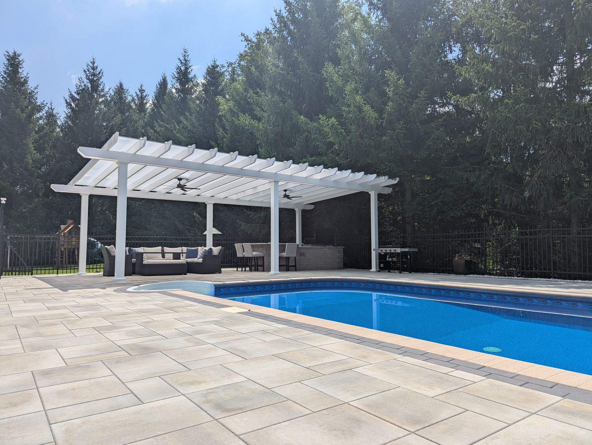A white pergola with seating and a pool area, surrounded by a paved patio and trees on a sunny day.