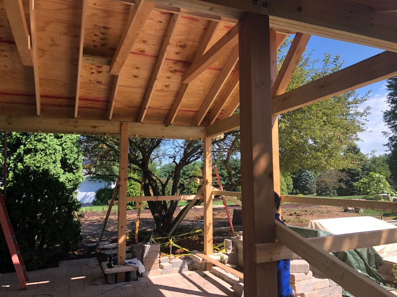 A wooden pavilion frame under construction, showing roof joists, support beams, and a view of trees and a yard.