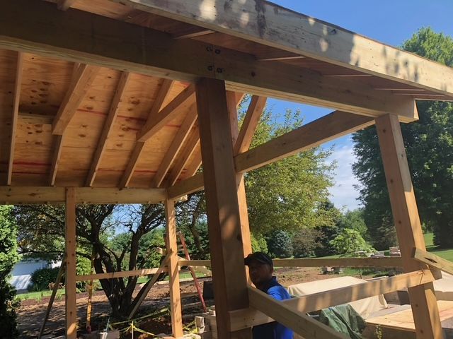 A person wearing a blue shirt works on the timber-frame construction of an open-air wooden structure outdoors.