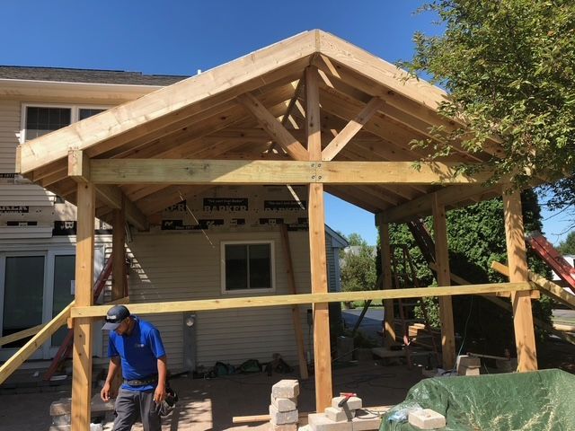 A worker stands in front of a house under construction with a newly built wooden porch frame attached.