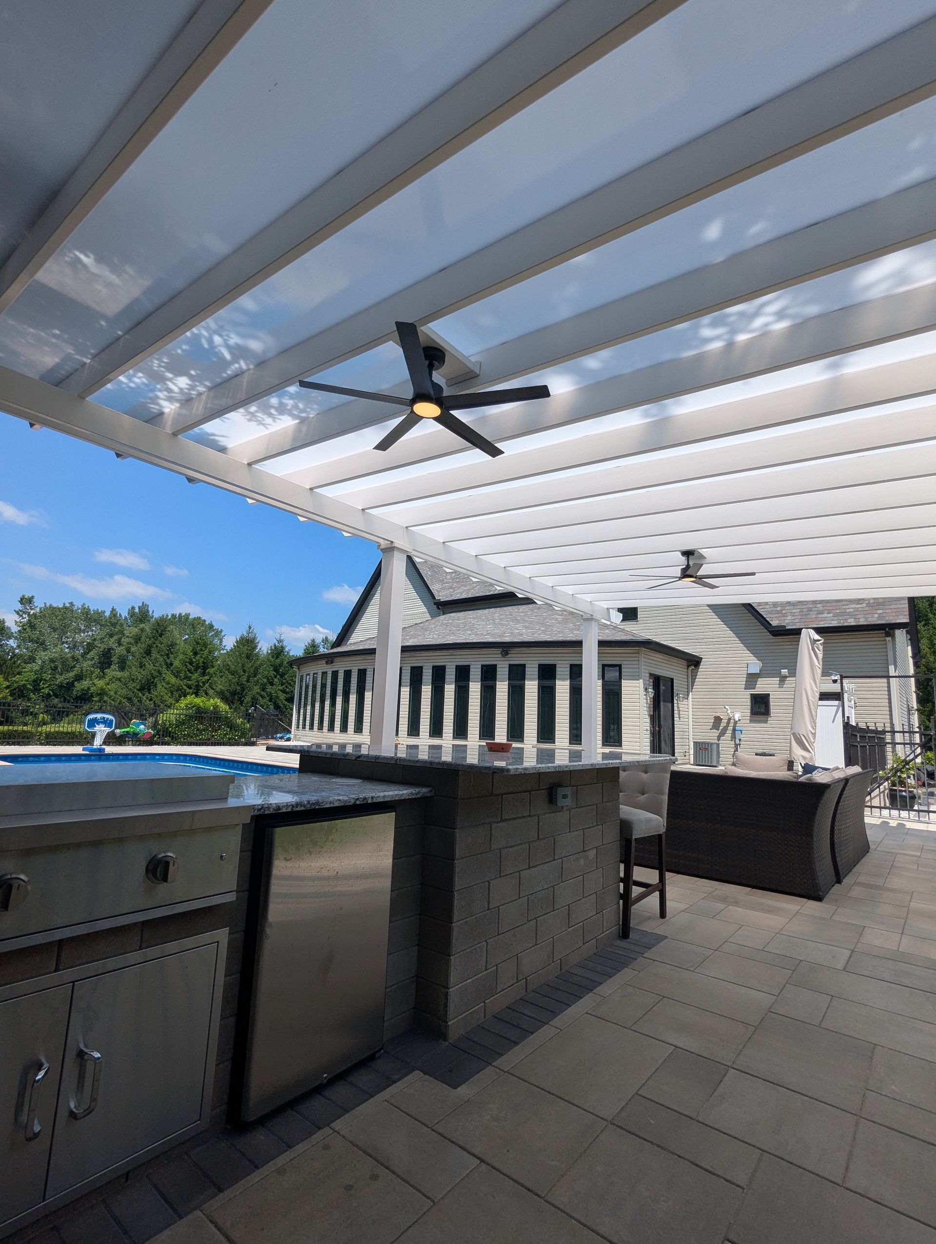 An outdoor patio featuring a covered bar area, a stainless steel grill, a ceiling fan, and a view of a nearby pool.