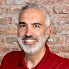 A man with a beard and a red shirt is smiling in front of a brick wall.