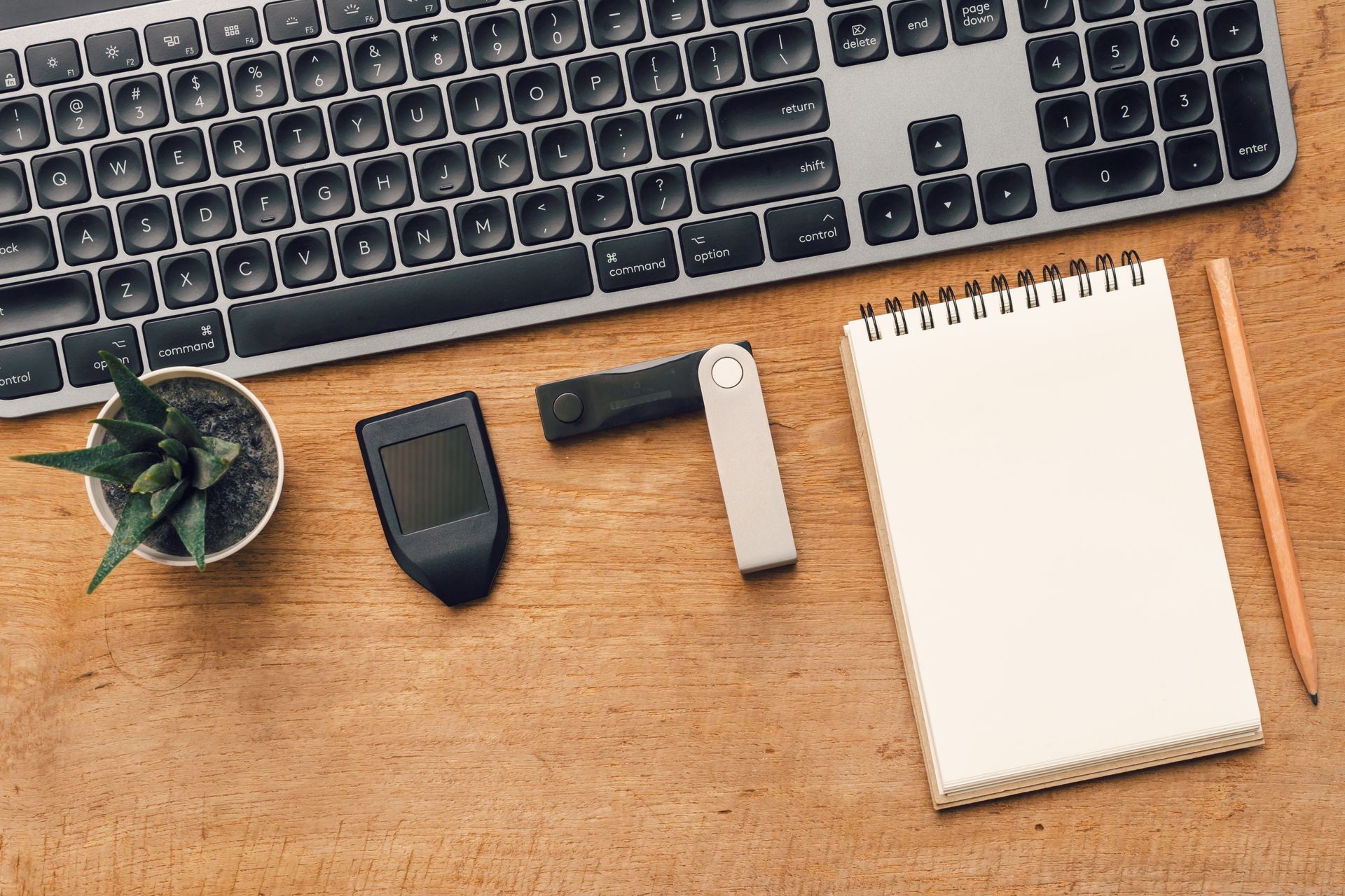 A wooden desk with a keyboard , notebook , pen and a plant.