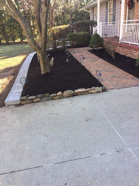 A brick walkway leads to a house porch next to a tree-lined flower bed with dark mulch and a stone border.