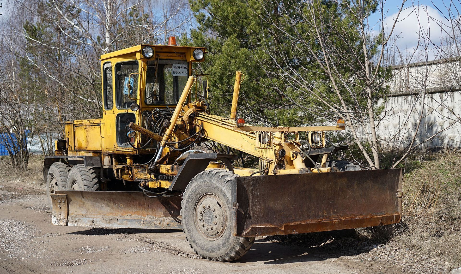 A yellow motor grader with a front-mounted blade parked on a dirt path with trees and a building in the background.