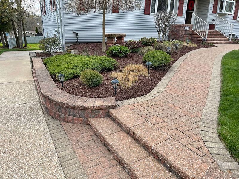 A brick-paved walkway with stairs leads to a house front, featuring a curved retaining wall and landscaped garden beds.
