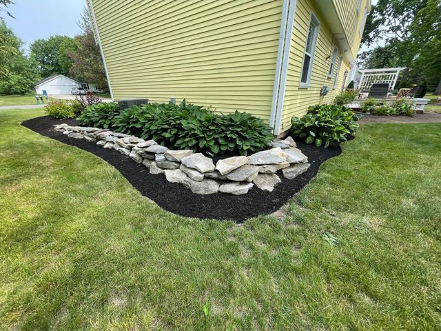 A yellow house with a stone retaining wall and lush green hostas surrounded by fresh black mulch in a grassy yard.