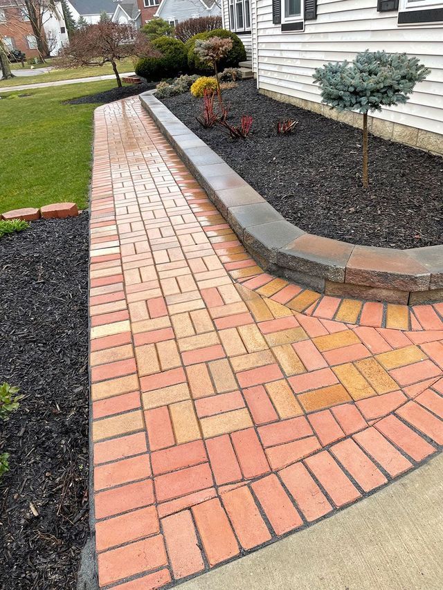 A red brick walkway with a herringbone pattern borders a raised stone retaining wall and black mulch flower bed.