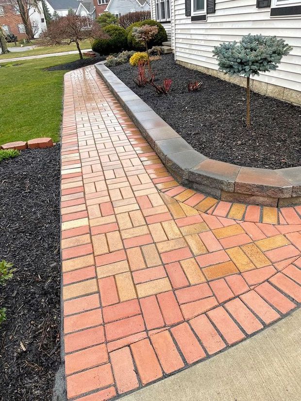 A red brick walkway with a herringbone pattern borders a raised stone retaining wall and black mulch flower bed.