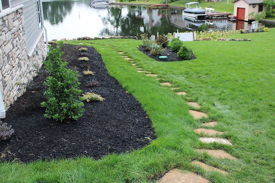 A stone path meanders through a green lawn toward a lake, alongside a house corner with a mulched garden bed.