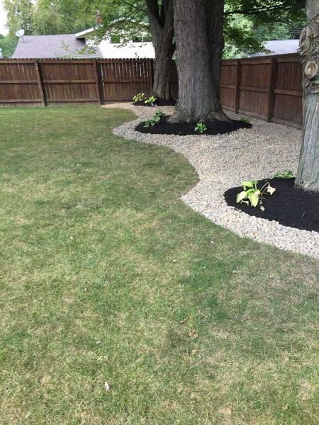 A backyard garden features a curved landscaping border filled with light gravel, mulch beds around trees, and a wood fence.