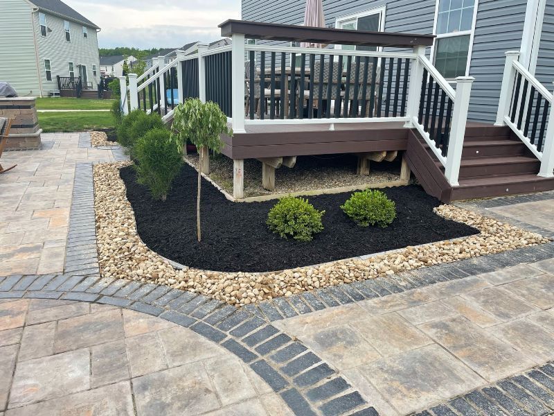 A raised deck with black railings above a landscaped garden bed containing small shrubs and trees, framed by stone pavers.