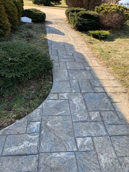 A stone-patterned concrete walkway winds through a yard with green bushes and patches of dry grass.