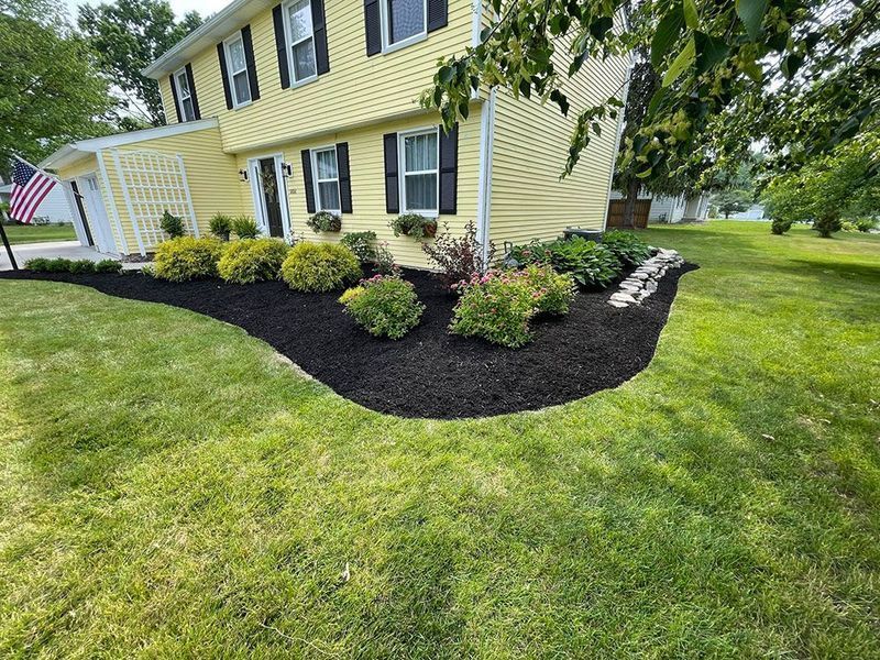 A yellow two-story house with a neat front flower bed filled with dark mulch, green shrubs, and small white rocks.