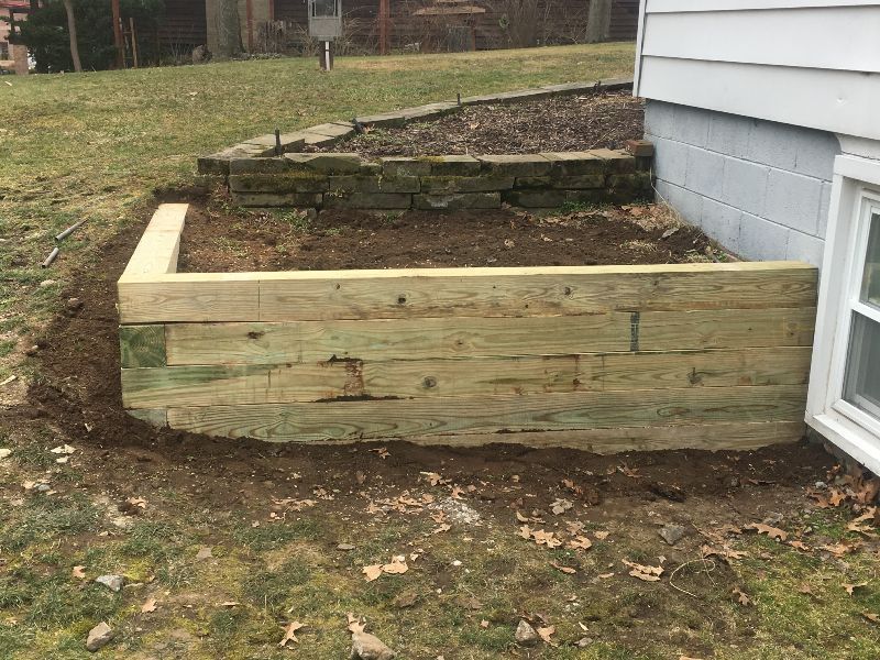 A freshly built timber retaining wall placed against the side of a house next to a lawn and an older stone wall.