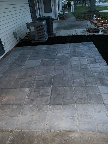 A new gray paver patio sits next to a house siding wall with an HVAC unit, bordering a lawn and walkway.