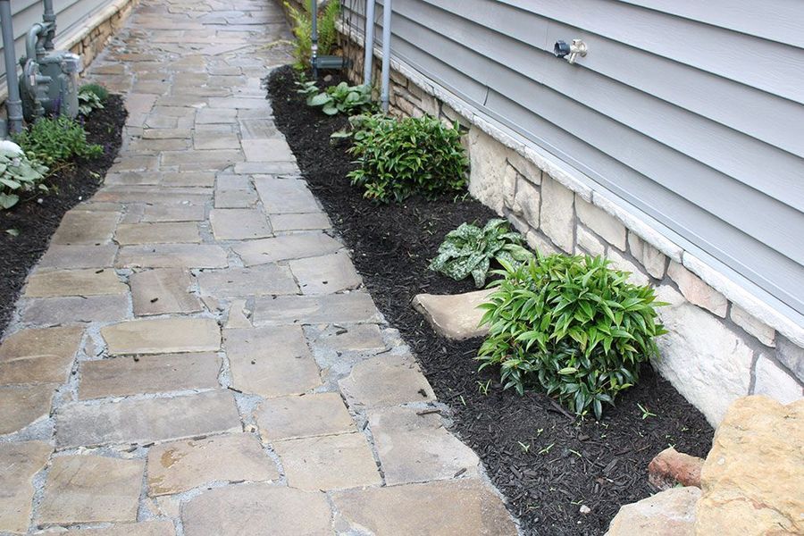 A stone walkway alongside a light-grey house, featuring a landscaped garden bed with mulch and small green shrubs.