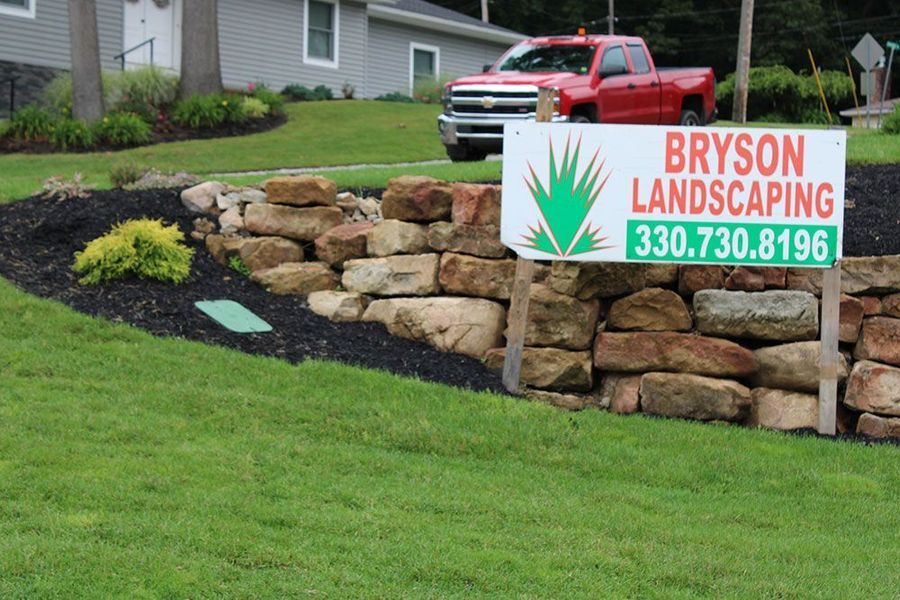 A stacked-stone retaining wall with a landscaped garden bed and a Bryson Landscaping sign on a residential lawn.