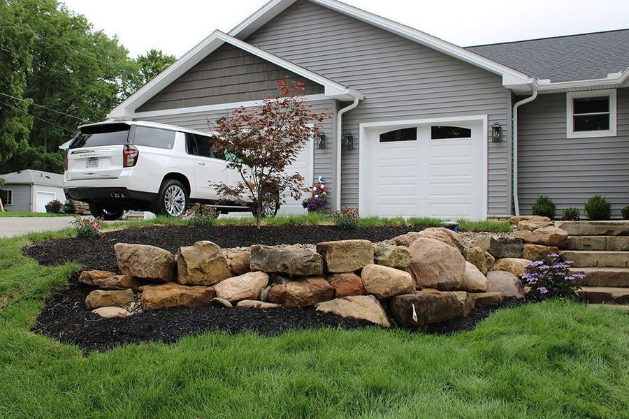 A gray house with a white SUV parked in the driveway behind a stone retaining wall and mulch bed.