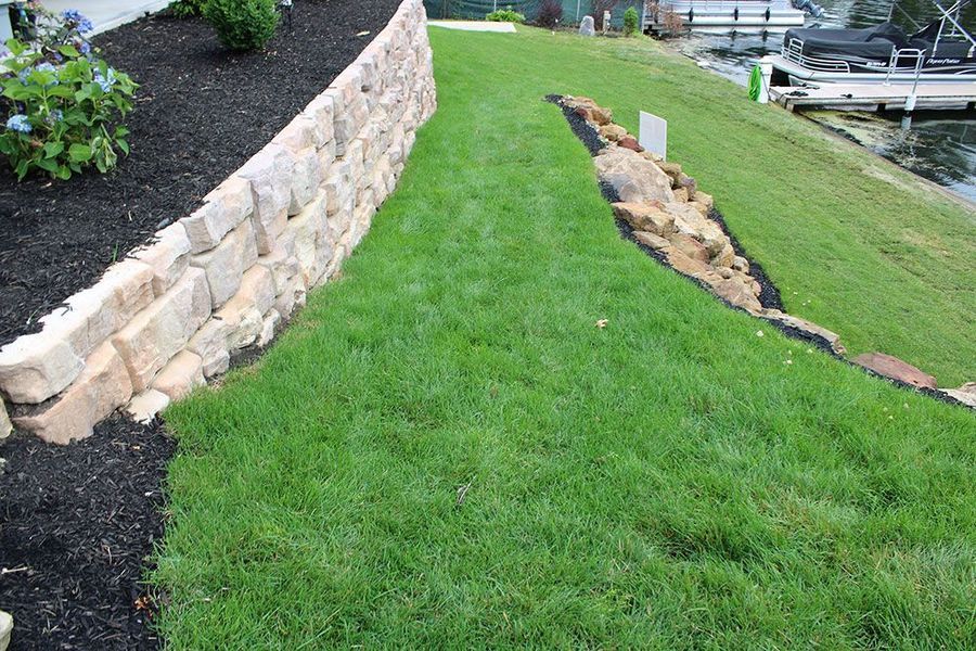A stone retaining wall next to a grassy lawn sloping down toward a lake with a boat docked at the water's edge.