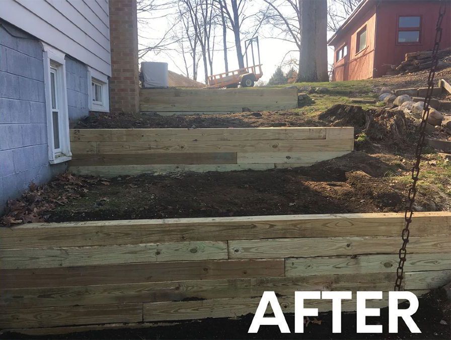 Three tiered wooden retaining walls built against the side of a house on a sloped grassy yard.