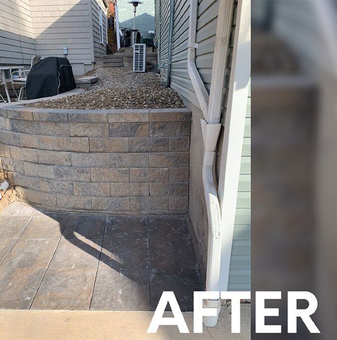 A stone retaining wall built next to a siding wall with a white downspout and a paved patio area.