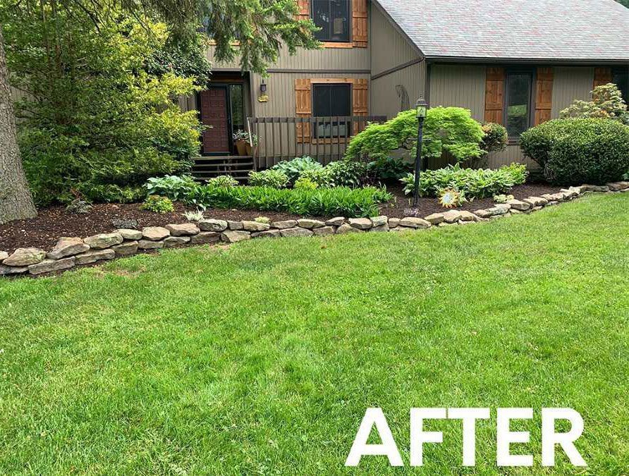 A well-maintained home garden with a stone border, lush green shrubs, and mulch against the exterior of a house.