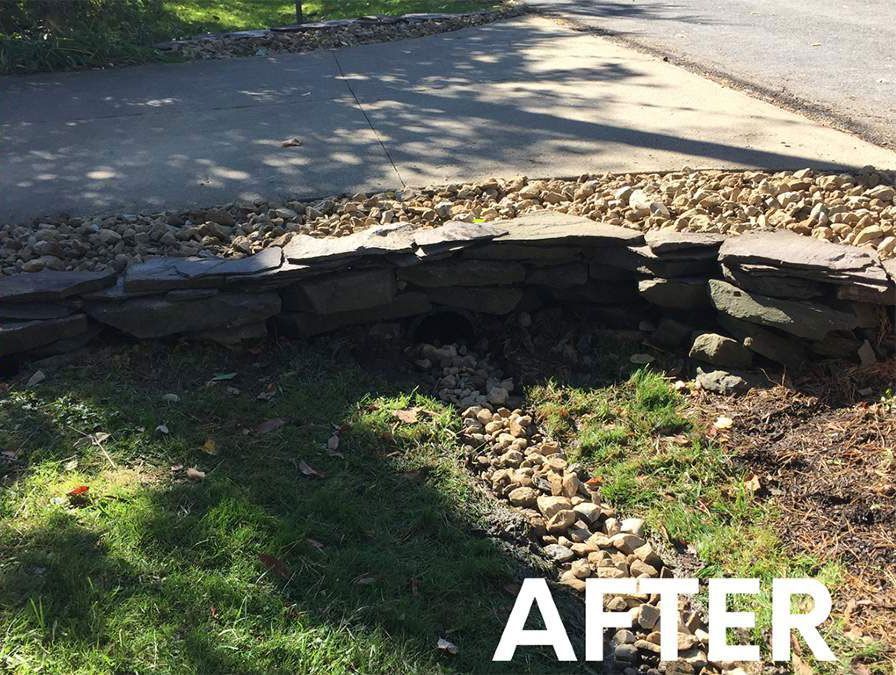 A stone retaining wall built over a small drainage ditch in a grassy yard, with a paved area in the background.